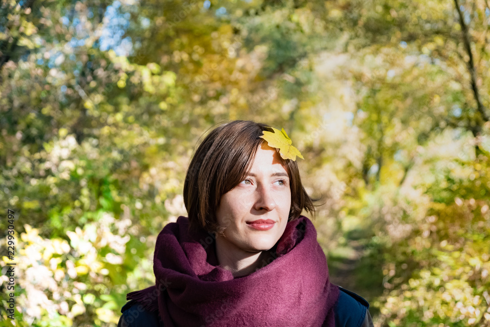 Portrait of young woman in scarf with autumn leave on her head. Maple leaf on the hair of a female person standing in beautiful green and yellow nature background Portrait of young woman in scarf with autumn leave on her head. Maple leaf on the hair of a female person standing in beautiful green and yellow nature background