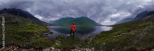 man stands by lake in red jacket in cloudy weather. cylindrical panorama 360vr