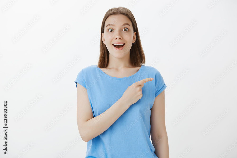 Excited attractive female friend with fair hair, staring with popped eyes at camera, smiling broadly while pointing at upper right corner, mesmerized with amazing copy space, standing over grey wall Excited attractive female friend with fair hair, staring with popped eyes at camera, smiling broadly while pointing at upper right corner, mesmerized with amazing copy space, standing over grey wall