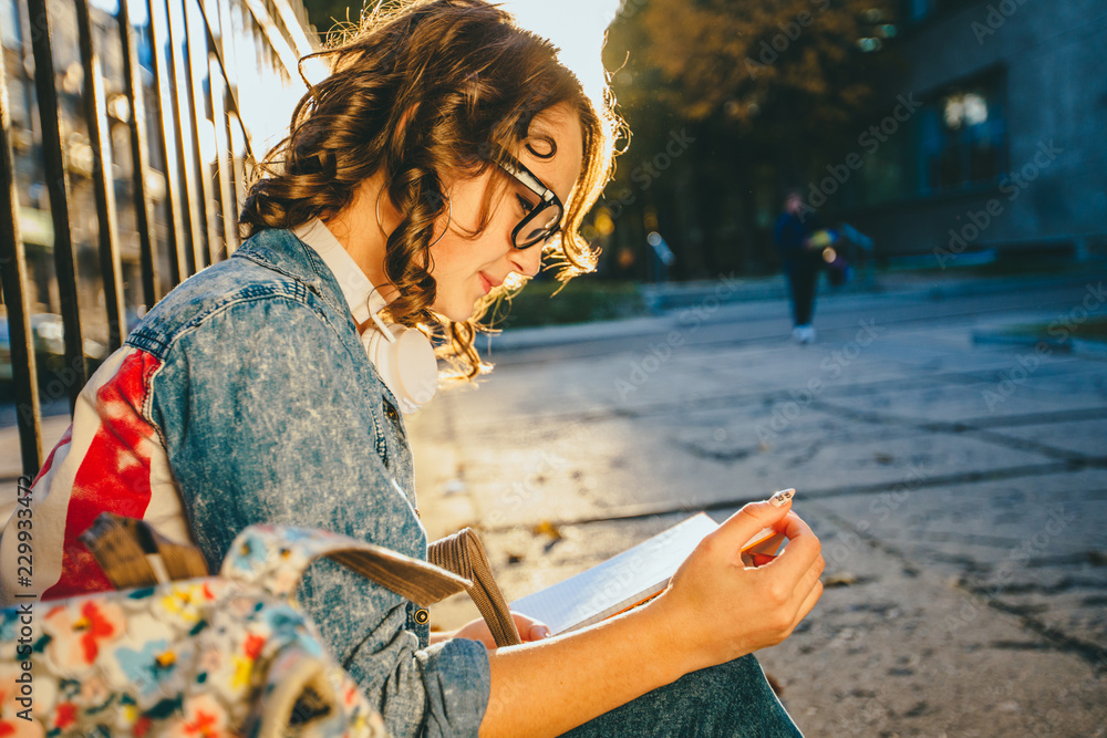 Side view portrait of young american female student girl in eyeglasses reading book outdoor in autumn city park in sunset.