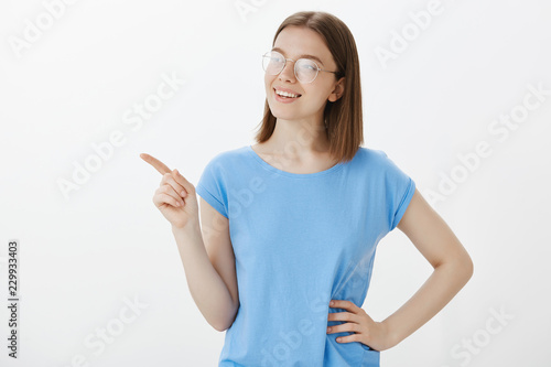 Studio shot of confident happy european female student in casual outfit, holding hand on hip, gazing and pointing left with index finger, smiling broadly, picking new furniture for apartment © Cookie Studio