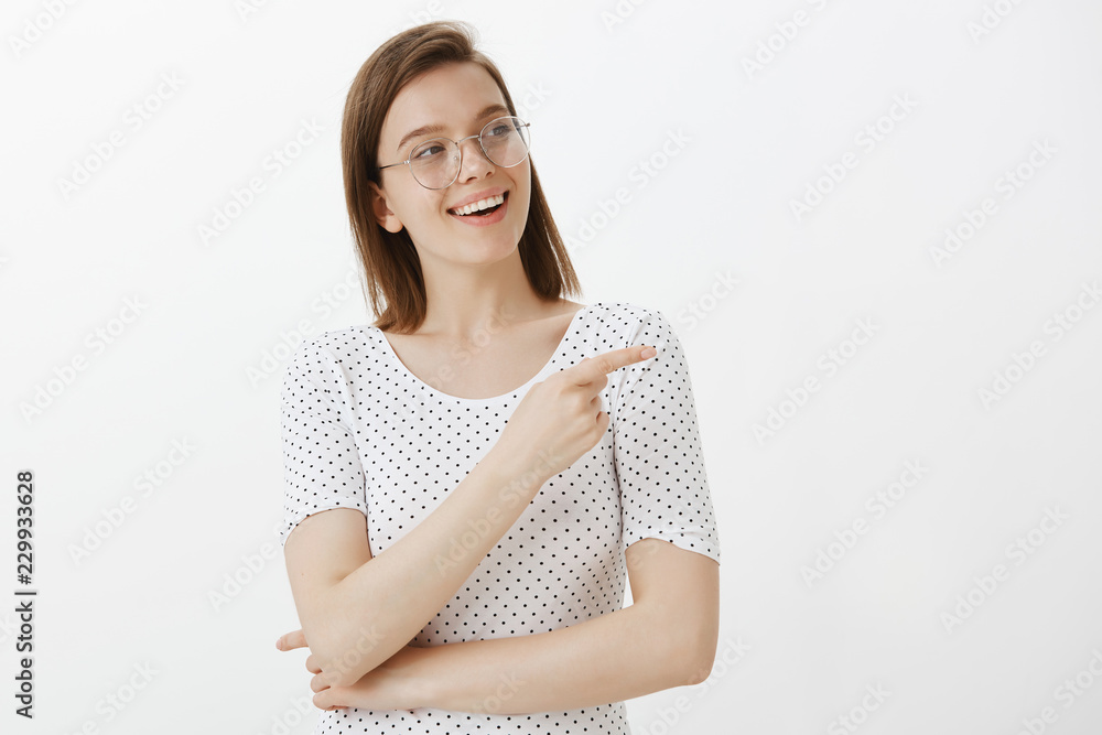 Waist-up shot of pleased and joyful good-looking woman in glasses, smiling broadly, gazing and pointing right with index finger, asking question about curious thing, standing over gray wall Waist-up shot of pleased and joyful good-looking woman in glasses, smiling broadly, gazing and pointing right with index finger, asking question about curious thing, standing over gray wall