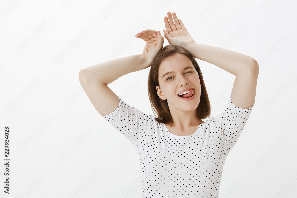 Sensual and flirty woman sticking out tongue and winking joyfully, smiling while showing bunny ears with hands over head, fooling around and having fun, enjoying youth over gray background Sensual and flirty woman sticking out tongue and winking joyfully, smiling while showing bunny ears with hands over head, fooling around and having fun, enjoying youth over gray background
