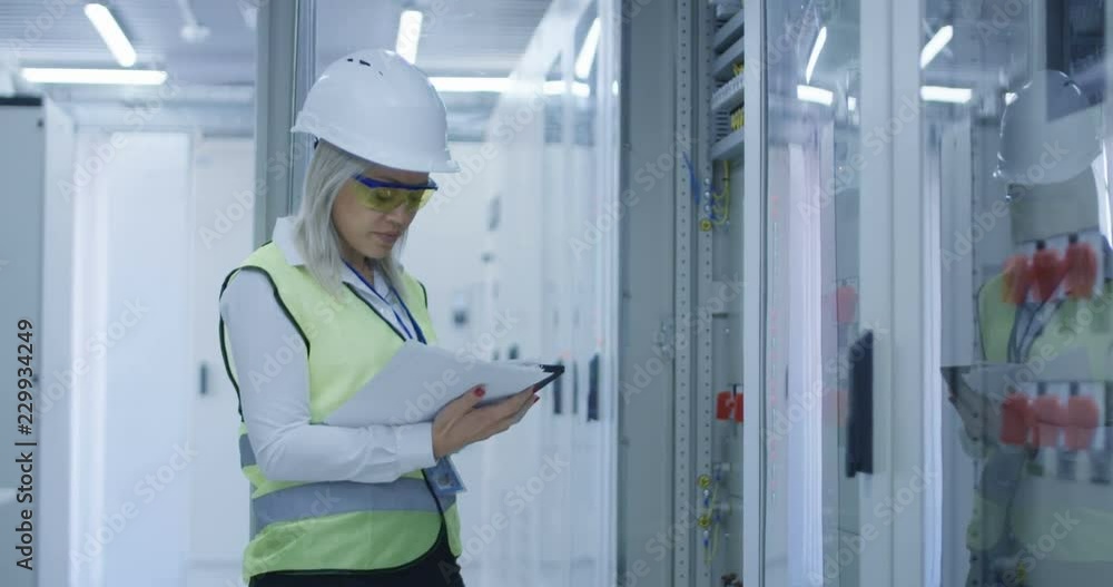 Formal blond woman in hardhat using tablet and checking electrical appliance in control center of solar plant Formal blond woman in hardhat using tablet and checking electrical appliance in control center of solar plant