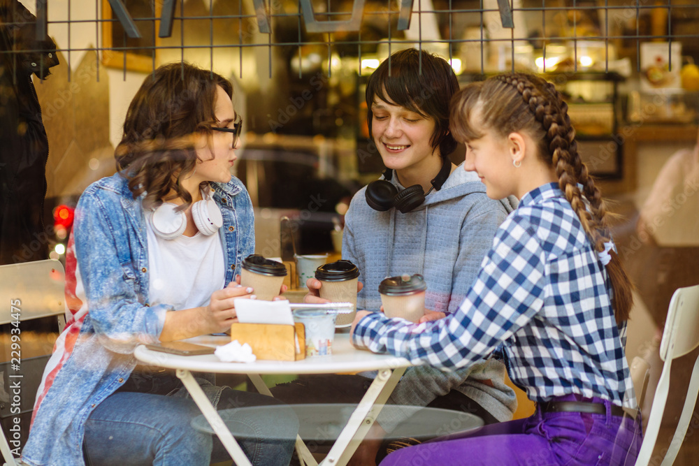Three happy young students friends sitting at table talking playing drinking coffee in modern coffee shop or tea house, shot through window glass. Three happy young students friends sitting at table talking playing drinking coffee in modern coffee shop or tea house, shot through window glass.