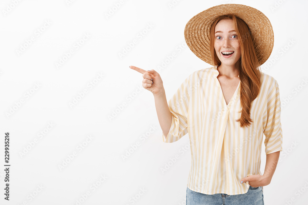 It is miracle look. Portrait of impressed and excited feminine timid redhead woman with freckles in straw hat and striped summer blouse pointing left with amazement smiling broadly from thrill