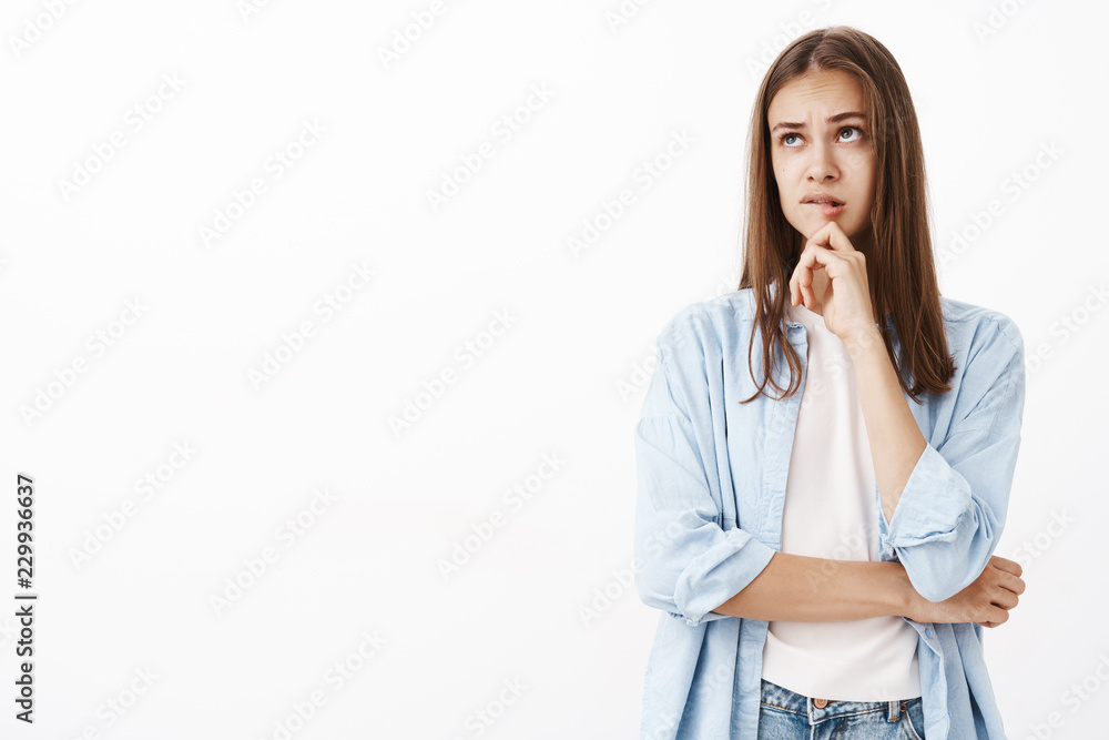 Girl trying make up solution thinking creating image in mind standing perplexed and thoughtful over gray background biting lower lip while thinking touching chin, looking up crossing arm over chest Girl trying make up solution thinking creating image in mind standing perplexed and thoughtful over gray background biting lower lip while thinking touching chin, looking up crossing arm over chest