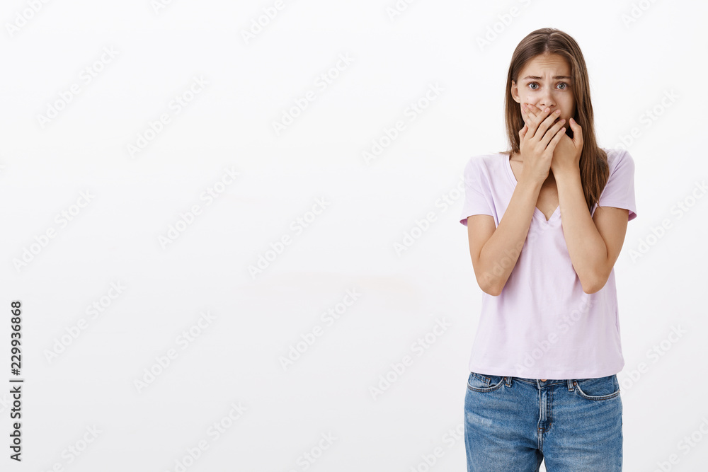Woman witnessing terrible accident feeling shocked and panicking. Portrait of terrified upset young european female covering mouth with both palms frowning screaming from fear over white wall Woman witnessing terrible accident feeling shocked and panicking. Portrait of terrified upset young european female covering mouth with both palms frowning screaming from fear over white wall