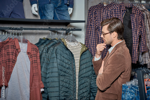 handsome young man with hand on chin looking at stylish clothes in boutique