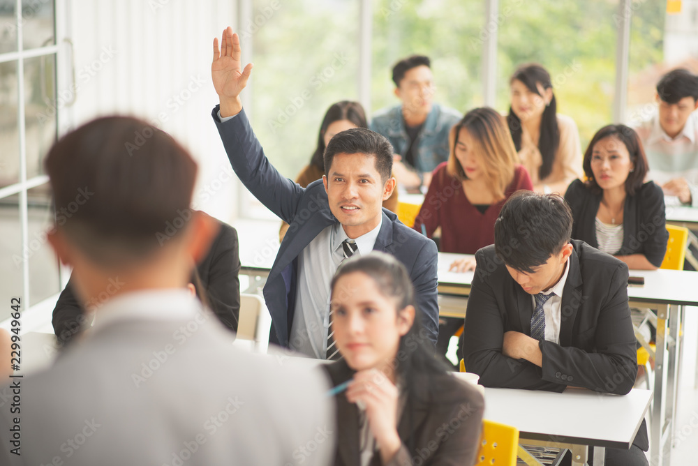 Man speaker talking in front of seminar meeting room with many audiences for business concept, some audiences raising up hand for asking in question and answer time in meeting room Man speaker talking in front of seminar meeting room with many audiences for business concept, some audiences raising up hand for asking in question and answer time in meeting room