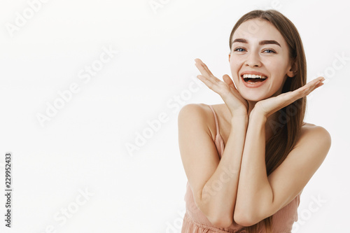 Waist-up shot of beautiful and healthy charming woman with natural beauty holding palms near jawline and smiling broadly pleased with perfect skin condition, posing cheerfully over gray background