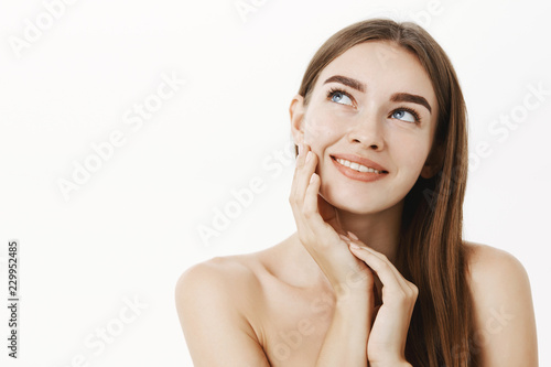 Woman applying cream on face feeling skin soft and tender standing dreamy and delighted with result gazing at upper left corner with sensual smile touching cheek posing naked over grey wall