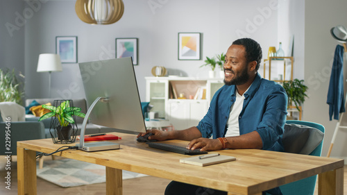 Happy Young Man Takes Place at His Desk and Starts Working on a Personal Computer. In the Background Cozy Living Room. © Gorodenkoff
