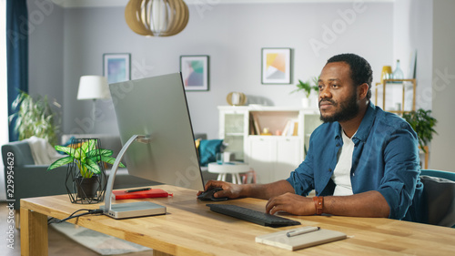 Tired African Amertican Man Works on a Personal Computer while Sitting at the Desk of His Cozy Living Room. © Gorodenkoff