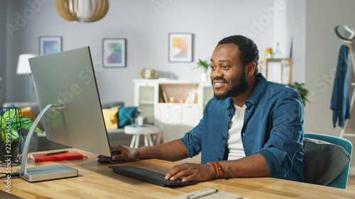 Handsome African Amertican Man Works on a Personal Computer while Sitting at the Desk of His Cozy Living Room.