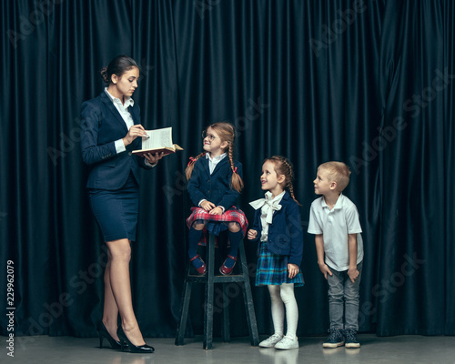 Cute smiling happy stylish children and female teacher on dark background. Beautiful stylish teen girls and boy standing together and posing on the school stage in front of the curtain. Classic style