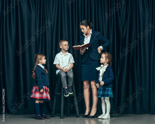 Cute smiling happy stylish children and female teacher on dark background. Beautiful stylish teen girls and boy standing together and posing on the school stage in front of the curtain. Classic style
