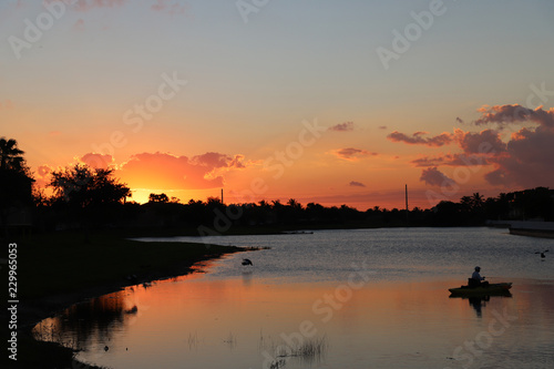 Mature man fishing from the boat on the pond at sunset, ducks swimming in foreground,