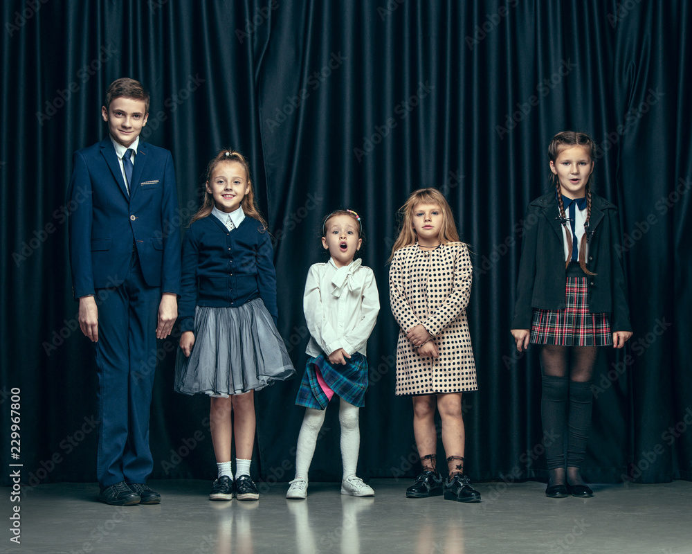 Cute surprised stylish children on dark background. Beautiful stylish teen  girls and boy standing together and posing on the school stage in front of  the curtain. Classic style. Kids fashion and Stock