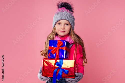 Happy little blonde curly hairstyle girl in knitted pink gray hat ,scarf and mittens with christmas gift box pink background studio. new Year present in hands of a female child making a wish copyspace