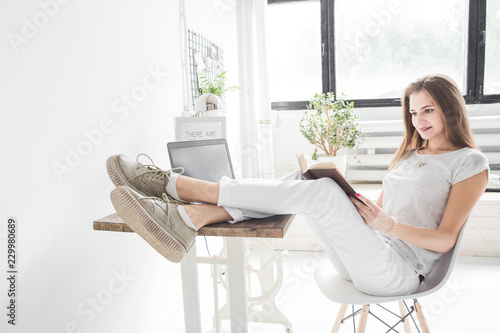 Young business woman working at home and reading a book with her legs on the table. Creative Scandinavian style workspace
