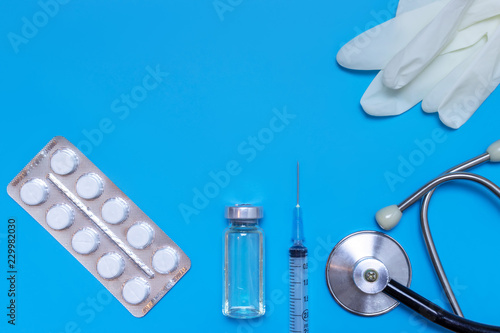 Layout of medical equipment, gloves, syringe, vial, stethoscope and tablets in blister on blue background with copy space, flat lay, copy space.