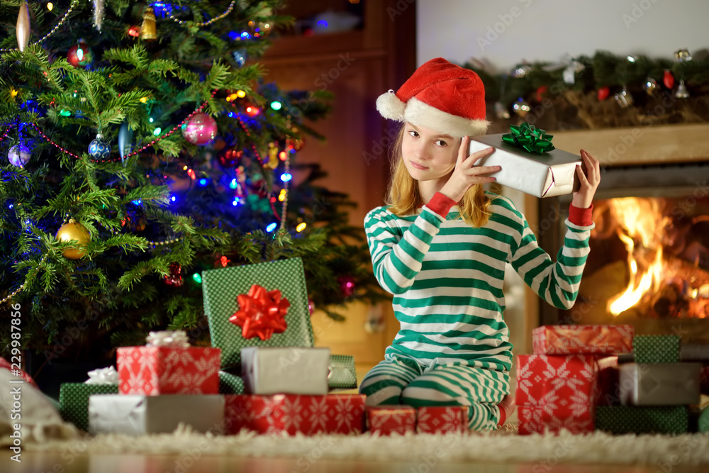 Happy little girl wearing Christmas pajamas playing by a fireplace in a cozy dark living room on Christmas eve. Happy little girl wearing Christmas pajamas playing by a fireplace in a cozy dark living room on Christmas eve.