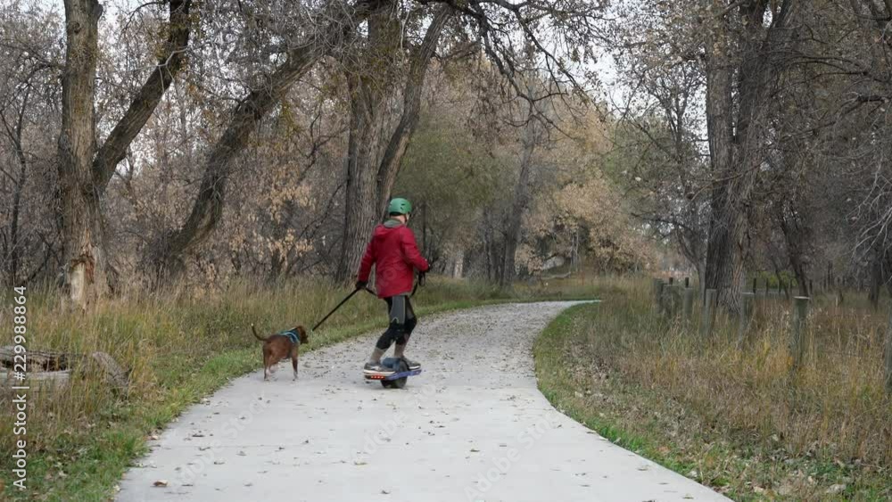 A senior man is slowly riding an electric skateboard with a dog on a leash - a paved trail with a fall scenery in northern Colorado A senior man is slowly riding an electric skateboard with a dog on a leash - a paved trail with a fall scenery in northern Colorado