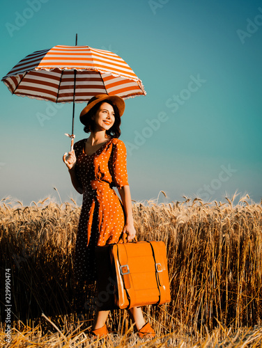 portrait of the beautiful young woman with red umbrella and brown suitcase standing on the field