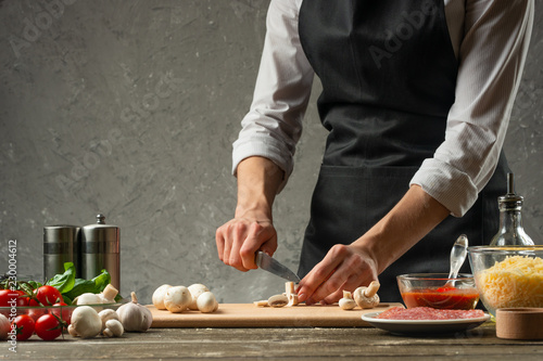 The concept of nutrition. The chef cuts mushrooms on the background of a concrete wall  with the ingredients for the preparation of pizza  pizza sauce  With empty space