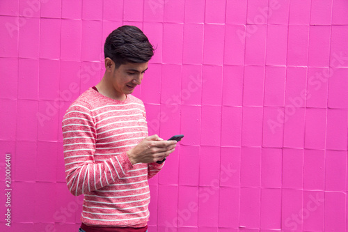 Photography A young handsome Latino guy, in a striped sweater and jeans, against a bright pink wall, looking at his phone, checking his messages, or texting, or surfing the web