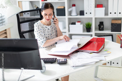 A young girl sitting in the office at the computer Desk, working with documents and talking on the phone.