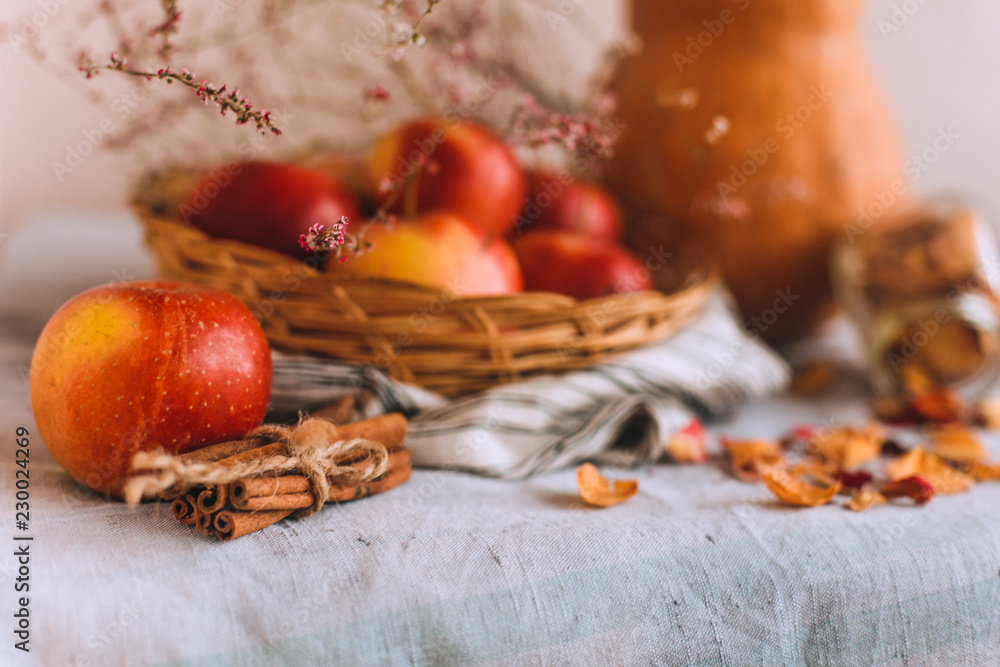 Still life with cinnamon sticks wrapped in twine, pumpkin, apples in a wicker basket stand on a striped linen cloth, walnuts and flower petals in bank. Concept of home comfort in autumn or winter. Still life with cinnamon sticks wrapped in twine, pumpkin, apples in a wicker basket stand on a striped linen cloth, walnuts and flower petals in bank. Concept of home comfort in autumn or winter.