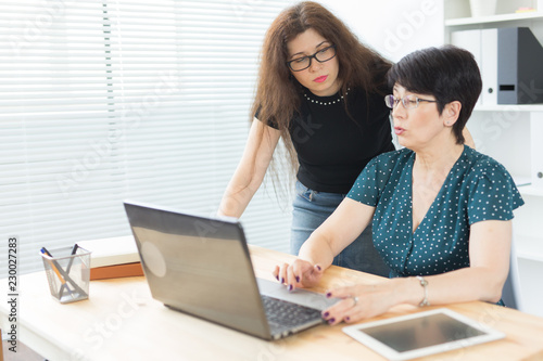 Technology, business and people concept - Young woman explaining something about laptop to her female colleague