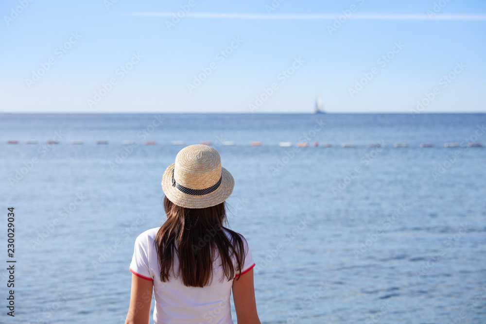 Beautiful girl looks at the sea. Young girl in a hat looking at a calm sea and blue skies back view.