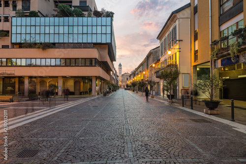 Fototapeta Naklejka Na Ścianę i Meble -  Characteristic street in the historic center of a city in northern Italy. Saronno (corso Italia), province of Varese, Lombardy at sunrise. At the bottom the Church of Saints Peter and Paul 