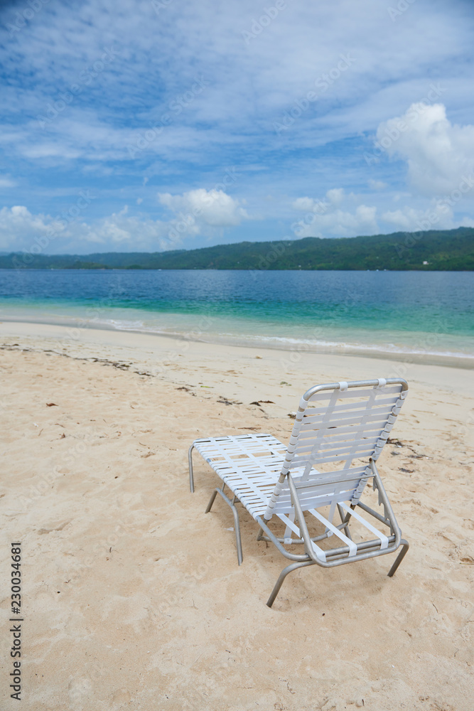 Small plastic deck chair standing on sand of Cayo Levantado beach against beautiful water of Atlantic Ocean in the Samana Bay. Lounge is white and lightwaight. water is blue and sky has some clouds. Small plastic deck chair standing on sand of Cayo Levantado beach against beautiful water of Atlantic Ocean in the Samana Bay. Lounge is white and lightwaight. water is blue and sky has some clouds.