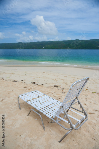 Small plastic deck chair standing on sand of Cayo Levantado beach against beautiful water of Atlantic Ocean in the Samana Bay. Lounge is white and lightwaight. water is blue and sky has some clouds. photo