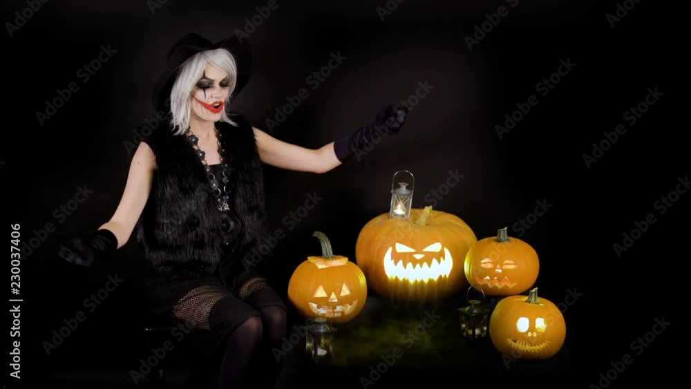 Scary beautiful girl witch laughs, taunts, gloats, celebrates halloween with funny glowing burning pumpkins in smoke. Woman with gray hair conjures, casts a spell Scary beautiful girl witch laughs, taunts, gloats, celebrates halloween with funny glowing burning pumpkins in smoke. Woman with gray hair conjures, casts a spell