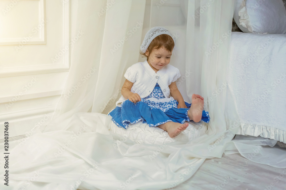 Funny and cute brunette little smiling girl playing and sitting relaxing in light white bedroom at home. White interior with big bed. Childhood, preschool, youth, relax concept Funny and cute brunette little smiling girl playing and sitting relaxing in light white bedroom at home. White interior with big bed. Childhood, preschool, youth, relax concept