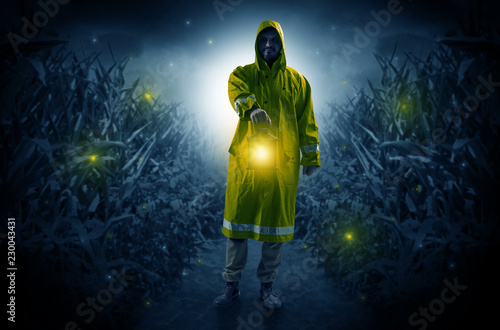 Man in raincoat at night coming from thicket and looking something with glowing lantern 