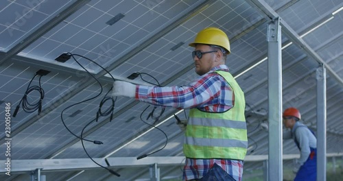 Medium shot of two electrical workers in hardhats connecting wires of solar panels at a solar farm