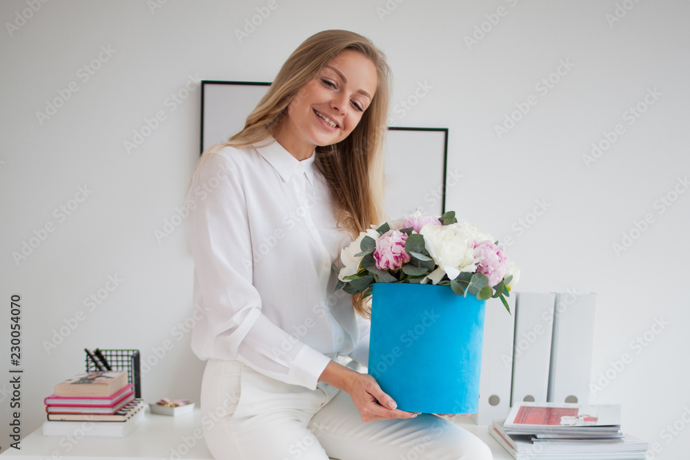 Stylish young blonde in office, white shirt and pants. Holds a luxurious bouquet of peonies in a blue hat box. Stylish young blonde in office, white shirt and pants. Holds a luxurious bouquet of peonies in a blue hat box.