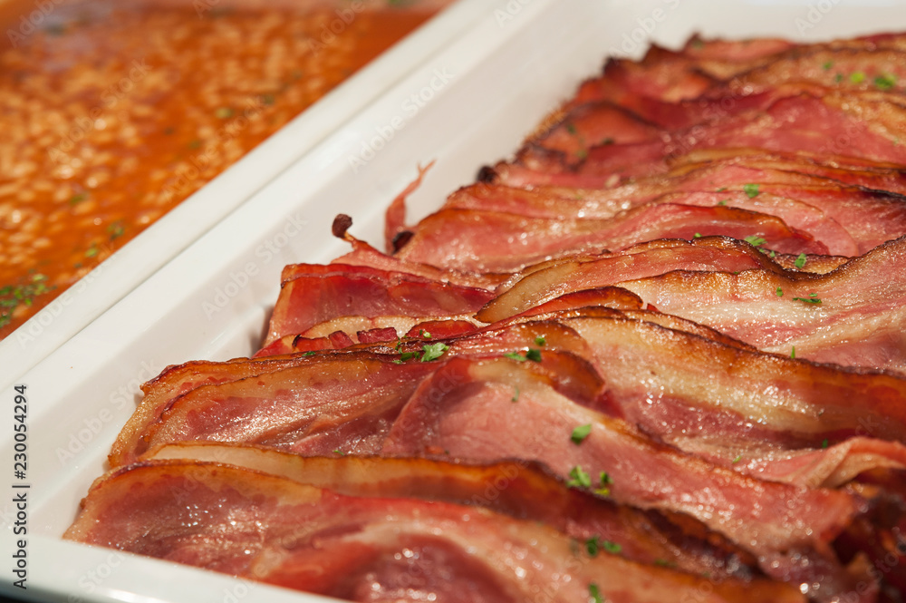 Closeup shot of trays of sliced grilled bacon and baked beans, healthy cooked breakfast options, adding protein and fiber to a diet. Horizontal shot with selective focus, concept for buffet-style meal Closeup shot of trays of sliced grilled bacon and baked beans, healthy cooked breakfast options, adding protein and fiber to a diet. Horizontal shot with selective focus, concept for buffet-style meal