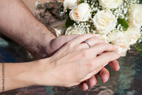 Close up shot of romantic couple, Caucasian female and male, delicately holding hands, with visible engagement ring on the woman's finger, and with an ivory roses wedding bouquet in the background