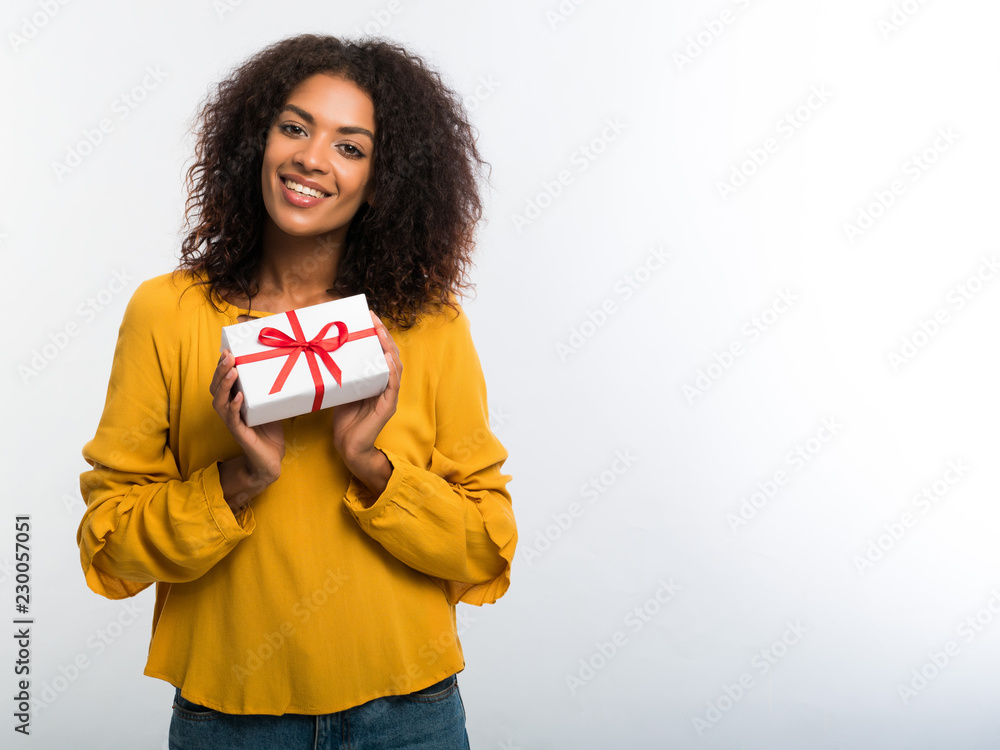 Cute african-american young woman in yellow autumn top holding gift box with red ribbon and bow on white wall background. Girl smiling, she is happy to get present Cute african-american young woman in yellow autumn top holding gift box with red ribbon and bow on white wall background. Girl smiling, she is happy to get present