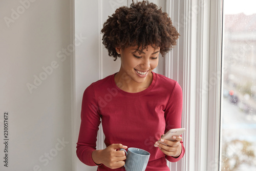 Happy pretty black girl happy to develop her own blog, rejoices having many followers, reads comments, drinks hot beverage, dressed in red clothes, stands near window. Hipster dials phone number photo