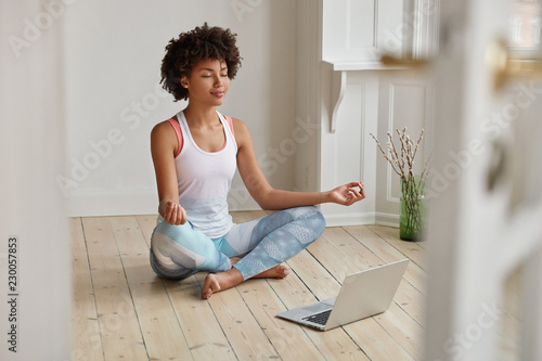 Fototapeta Relaxed dark skinned woman with sporty body, sits in zen pose, keeps legs crossed, watches yoga lessons on laptop computer using internet, meditates on floor in empty room