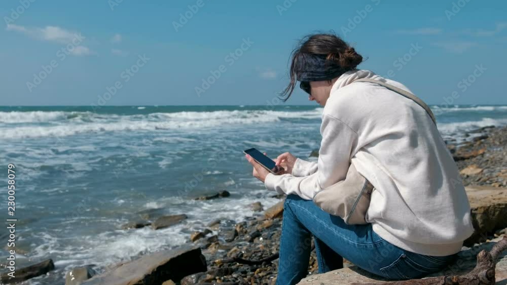 Woman plays the game on phone sitting on the sea shore in autumn. Woman plays the game on phone sitting on the sea shore in autumn.