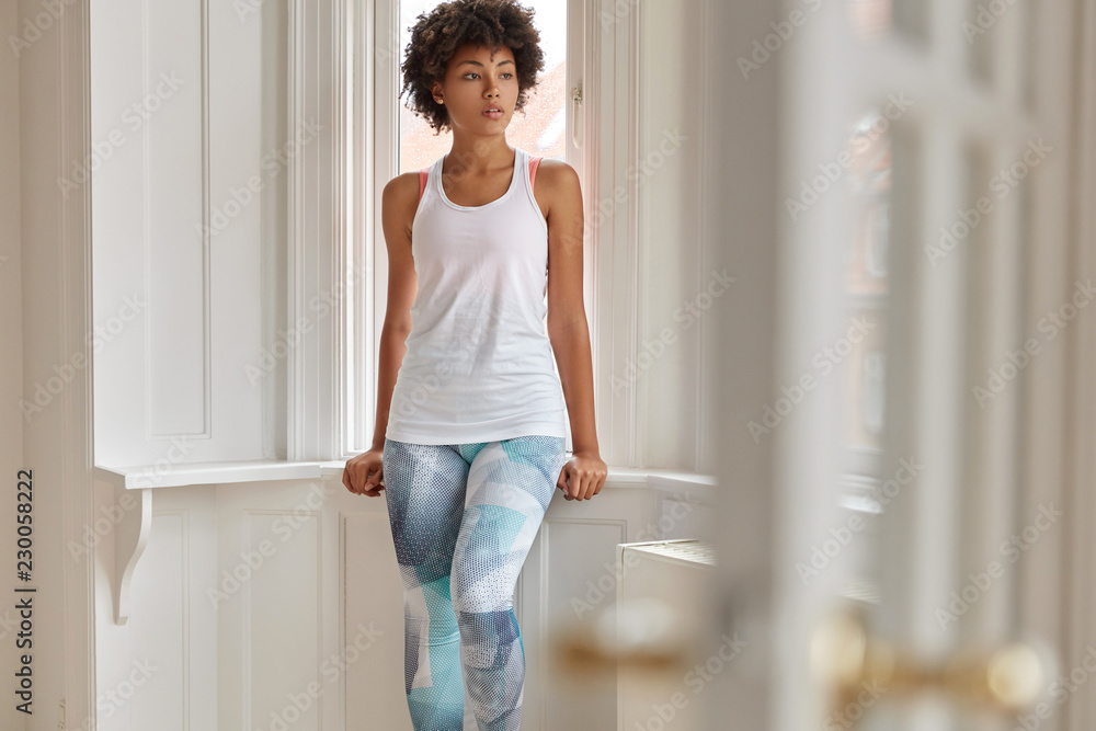 Fototapeta premium Photo of pretty relaxed fitness trianer rest at home after training, wears casual white vest and colorful leggings, leans at window sill, poses at balcony, has thoughtful expression. People, lifestyle